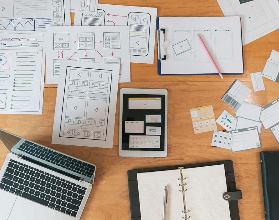 A desk with sketches, a laptop, and printed wireframes for an app